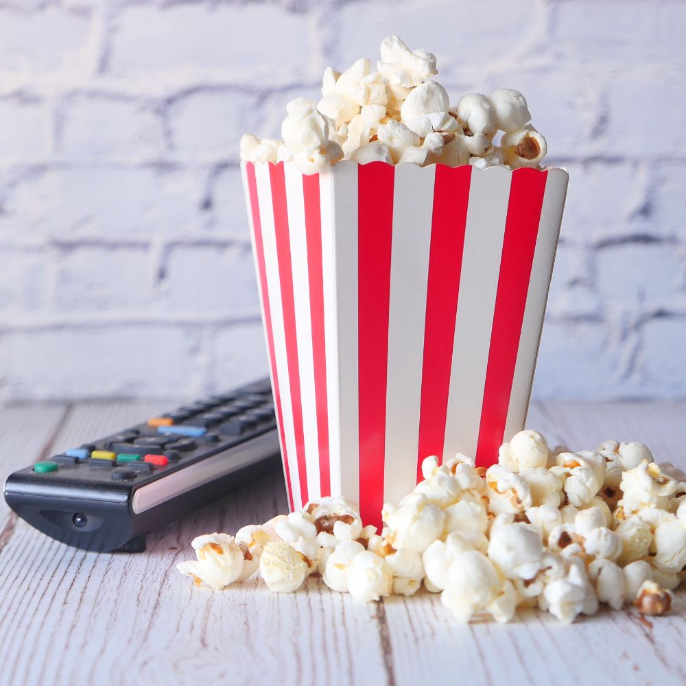 Premium IPTV Subscription | Red and white striped popcorn container filled with fresh popcorn next to a TV remote control on white wooden surface, representing home entertainment streaming experience